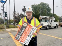 A man in a fluorescent yellow safety vest holds a sign advertising free soil and lead testing as he walks on a crosswalk. A utility truck idles at a red light.