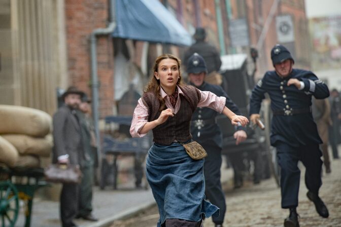 Millie Bobby Brown is in the center of the image mid run. She wears a faded blue skirt, a brown button up vest, and a light pink collared shirt underneath. The shirt sleeves are rolled up to her elbows and the skirt is gathered at the waist to shorten the hem length. Around her waist is also a small yellow pouch that peeks out from her vest. Brown's left arm is in front of her, slightly blocking her chest, while her right arm stretches behind her. Her arms are pumping alongside her as she sprints through a dirt street. Behind her, seemingly chasing after her, are two police officers. Their uniforms are from the 1800s (the time period of the film), with navy blue coats that hit mid-thigh with brass round buttons up the middle, and matching navy blue pants. The two officers also wear helmets, almost egg-shaped, with straps across their chins to help keep them in place. One of the officers on the left is partially blocked by Brown's frame, while the other officer on the right can be fully seen, his left hand holding a cylindrical tan item. In the background along the street are men in suits watching next to carts full of burlap sacks. A horse drawn carriage can be seen behind the two officers as well as signs for storefronts to the right, indicating a bustling city. 