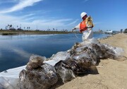 Plastic bags full of oil debris sit on the shore of a channel with a worker in white coveralls and an orange life preserver coils yellow rope.