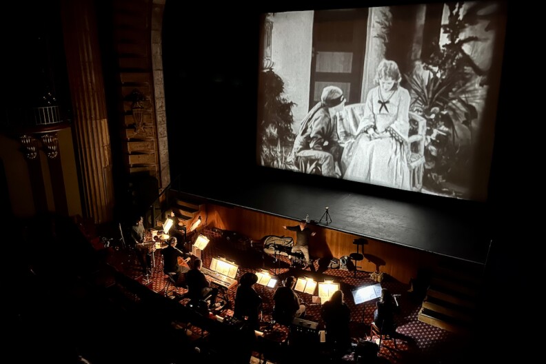 A small ensemble performs at the foot of a screen that is showing a black-and-white film