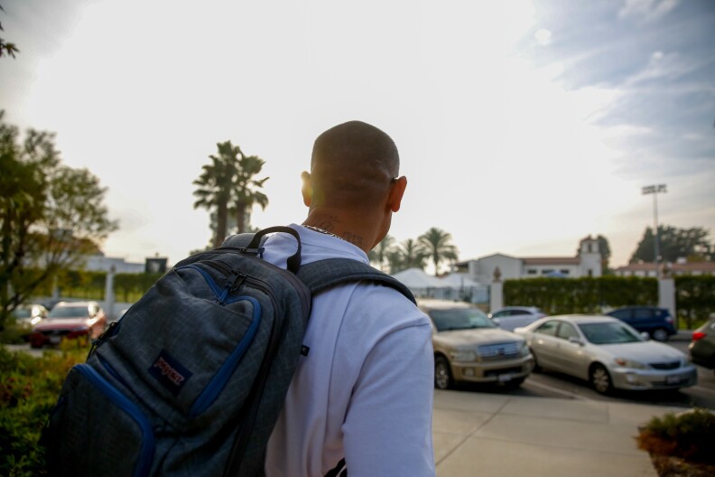 A photo captures a man wearing a white t-shirt and a backpack from behind walking toward a parking lot.