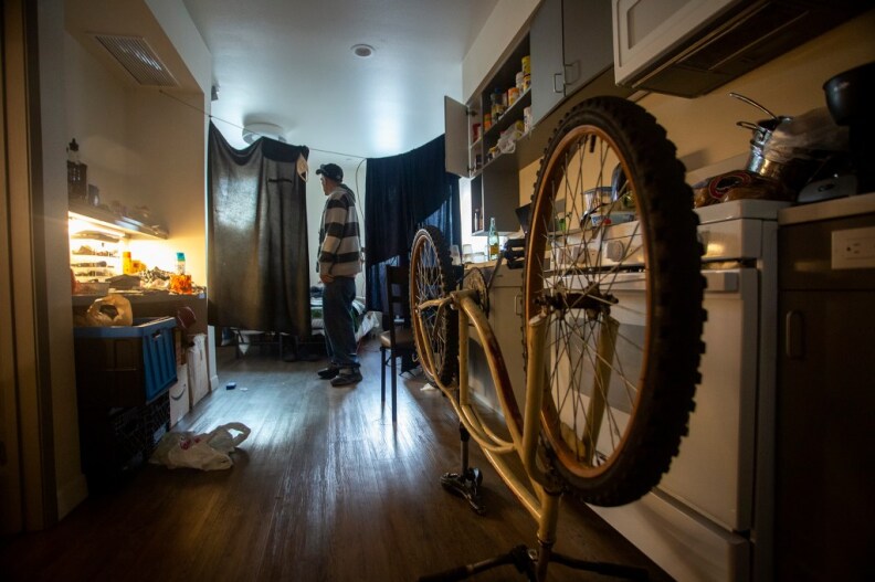 A man standing on the other side of the room looking at hanging sheets. A biking is in the foreground upside down in the kitchen.