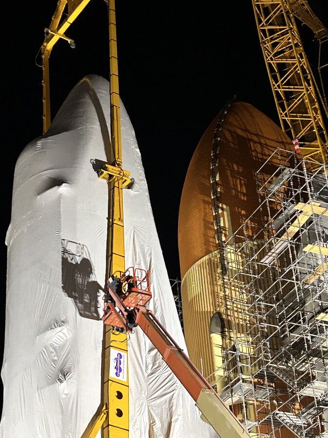 A close-up of the top of a white space shuttle orbiter behind held in the air by a towering yellow crane. The orbiter is relatively close to a vertical orange object that is roughly the same size. 