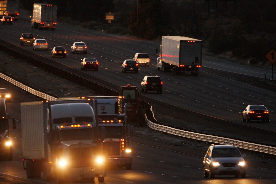 The 10 freeway in California at dusk, as heavy duty trucks and cars travel on both sides, their headlights and backlights glowing in the twilight