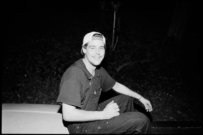 A black and white photograph of a man in a backwards hat and overalls.