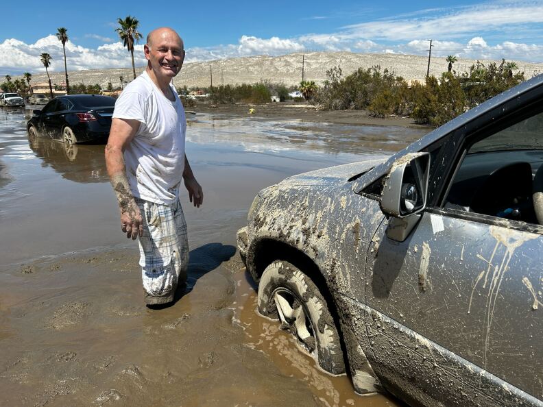 A middle aged, balding, light-skinned man wearing a white t-shirt and mud-splatted white pants stands knee-deep in mud beside a car stuck in the mud.