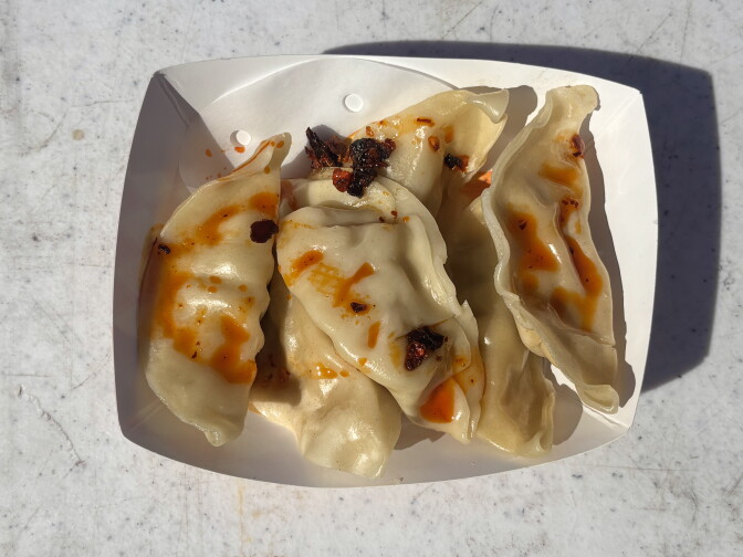 Overhead view of six boiled half-moon dumplings in a white paper boat on a weathered outdoor table. The dumplings are drizzled with orange chili oil and topped with clumps of dark, crispy fried chili.
