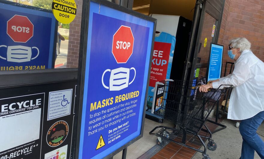 A large red and blue sign outside a grocery store shows a stop sign, a face mask, and large block letters reading "masks required." An older woman walks into the store wearing a face mask, short white hair, a white dress shirt and jeans.