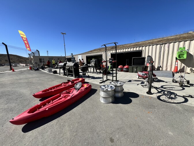 A variety of objects are on display in front of a storage-container like structure that's working as a walk-up shop. The objects include two bright red kayaks, grills, exercise equipment, and bicycles of all shapes and sizes. A large yellow and red flag sign attached to another building on the far left of the photo says "Now Open" on a bright, clear morning. 