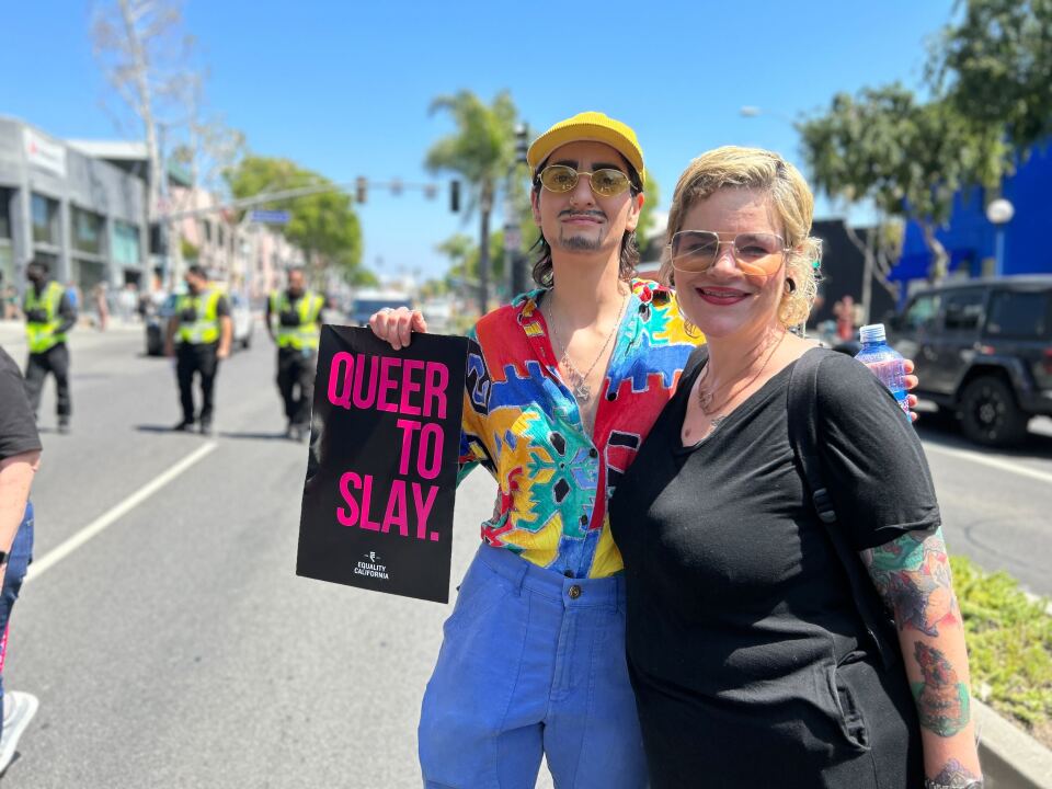 Two people with light skin tones smile for the camera on the street. The person on the left is in drag with a mustache and is holding a sign that says "Queer to slay." The person on the right has blonde hair and black clothing.
