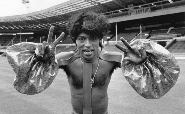 Little Richard in a costume featuring puffy sleeves, a strap up his chest, and appearing mostly shirtless. He gives peace signs with both hands from the field of a massive stadium.