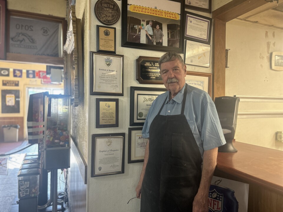 An older man with light skin and a grey mustache and short grey hair wears a black apron over a blue collared shirt. He stands against a wall with framed photos and plaques. 