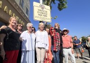 A crowd of people of varying ages are smiling and posing in front of a city street sign that reads "Tritch Family Square" and "Meeting the hardware needs of the Eagle Rock community since 1945" on a cloudless, sunny day.
