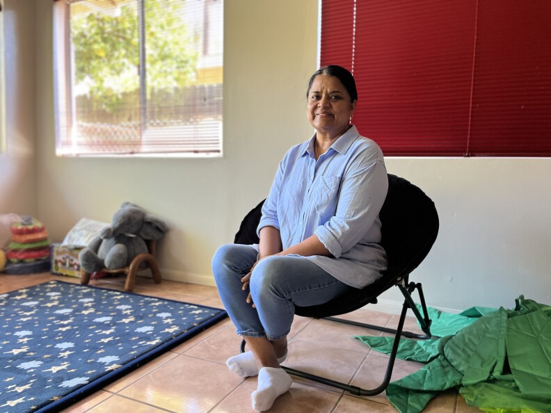 A woman with medium dark skin tone and blue shirt sits in a chair in a room full of children's toys. 