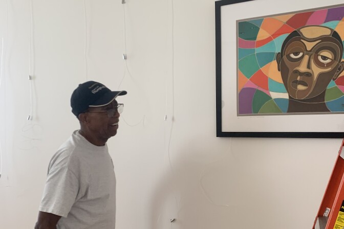 A man with dark skin and wearing hat and glasses looks at a painting of a man with a similar skin tone, with an impressionistic style.