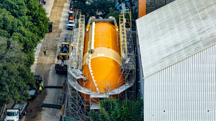 An enormous bright orange tank is surrounded by construction scaffolding. The tank is sitting next to a building outside, dwarfing the construction vehicles and cars parked next to it.