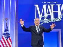A man wearing a dark suit and tie holds his arms out. His mouth is open. Behind him is a sign against a blue background that reads, "MAHA Summit" and an American flag stands to his right