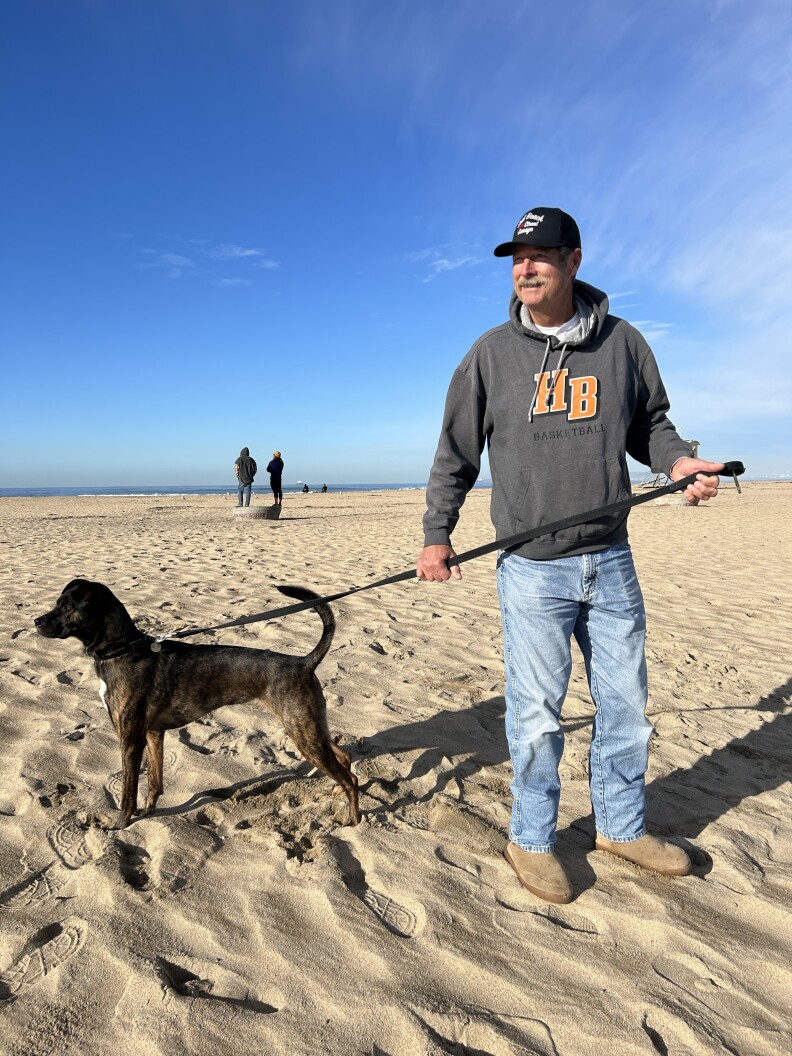A white man with a graying mustache wears a baseball cap, jeans and a gray sweatshirt on a wide, sunny beach. He's holding a leash with a medium sized black-brown dog at the end, looking off camera. 