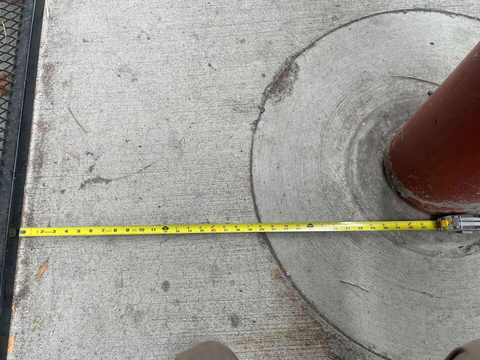  A measuring tape is placed between two obstructions on the sidewalk, a black piece of fencing and a red column. The tape shows that there are approximately 30-31 inches of space between the two obstacles.