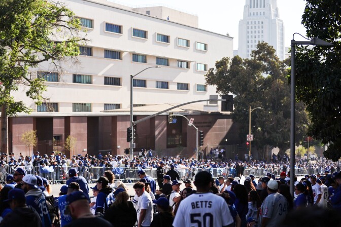A large group of people wearing Dodgers attire stand at both sides of a street behind metal barricades. 