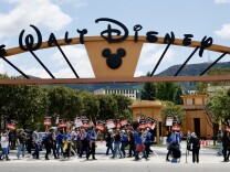 Picketers, many of them wearing blue T-shirts, walk back and forth in front of a yellow building under a huge sign reading "Walt Disney" in black lettering. They're holding red and black signs with white lettering, with the word "Strike!" in large, clear print under more writing that's too small to make out.