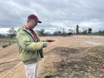A middle aged man with light skin wears a light green jacket and red baseball cap and gestures toward the ground while standing on a dirt lot on a rainy day. 