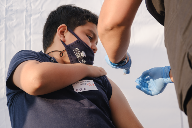 A boy in a navy blue shirt holds up his sleeve while his arm is cleaned in preparation for a coronavirus vaccine. 