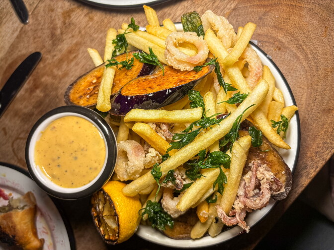 An overhead shot of a plate piled with fried calamari, eggplant, and fries, garnished with fried parsley, served with a small bowl of orange aioli and a charred lemon half.
