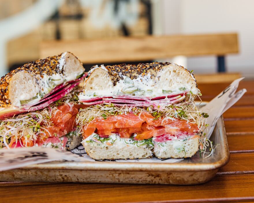 An everything bagel is cut in half, showing the luscious insides; layers of cream cheese, smoked salmon, fresh herbs. It's sitting on a metal tray on a wooden table.
