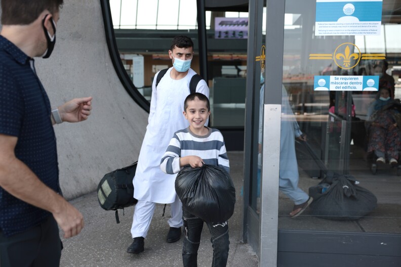 An Afghan boy, around age 9, in a striped shirt walks out of Dulles airport with a masked man following behind him, presumably his father.