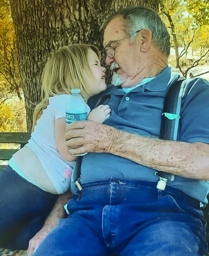 A girl approximately age four is shown hugging and making a face at an older man, her grandfather. They sit on a bench in front of a tree. The girl has blond hair and wears a white t-shirt and blue leggings. The man wears jeans, suspenders and a faded blue polo shirt. He has gray hair and a gray mustache and goatee. 