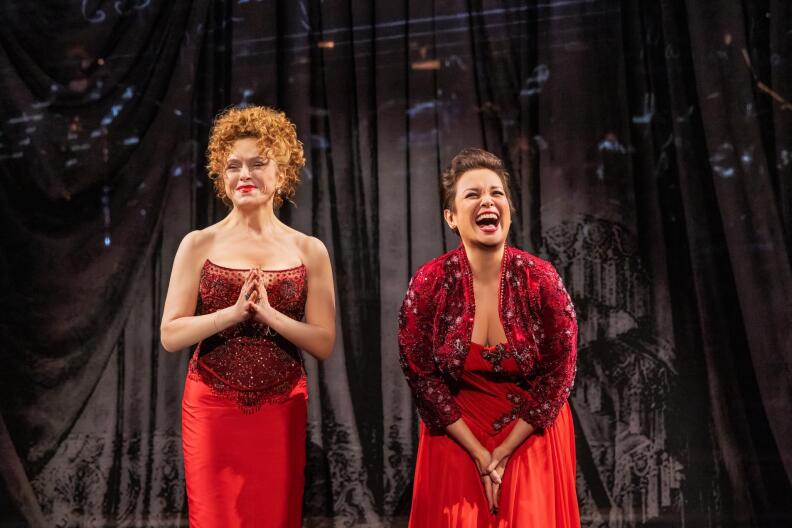 Broadway stars Bernadette Peters and Lea Salonga are both smiling and laughing on stage, dressed in red, with a sheer black curtain behind them.