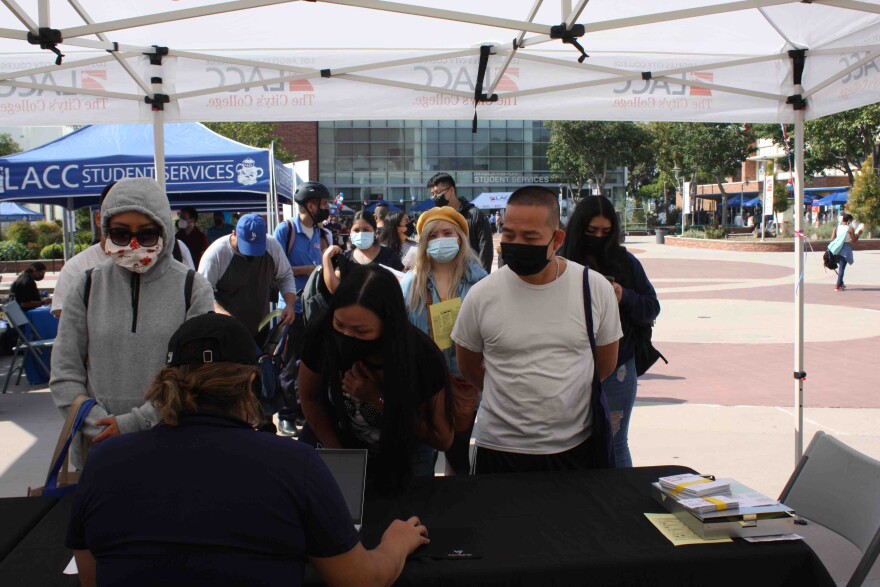 Students wearing masks stand in a long line waiting to see a school official seated in a pop-up tent outdoors at Los Angeles City College. In the background is another pop-up tent with a blue canopy that says LACC Student Services.
