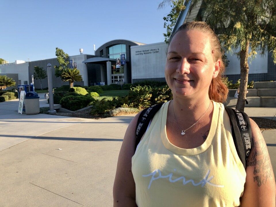 A light-skinned woman in a bright yellow tank top and a backpack smiles at the camera. In the background is a large college building.