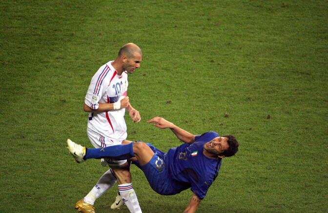 A soccer player in a white uniform stands as he gestures watching another player in a blue uniform as he falls to the ground on a field.