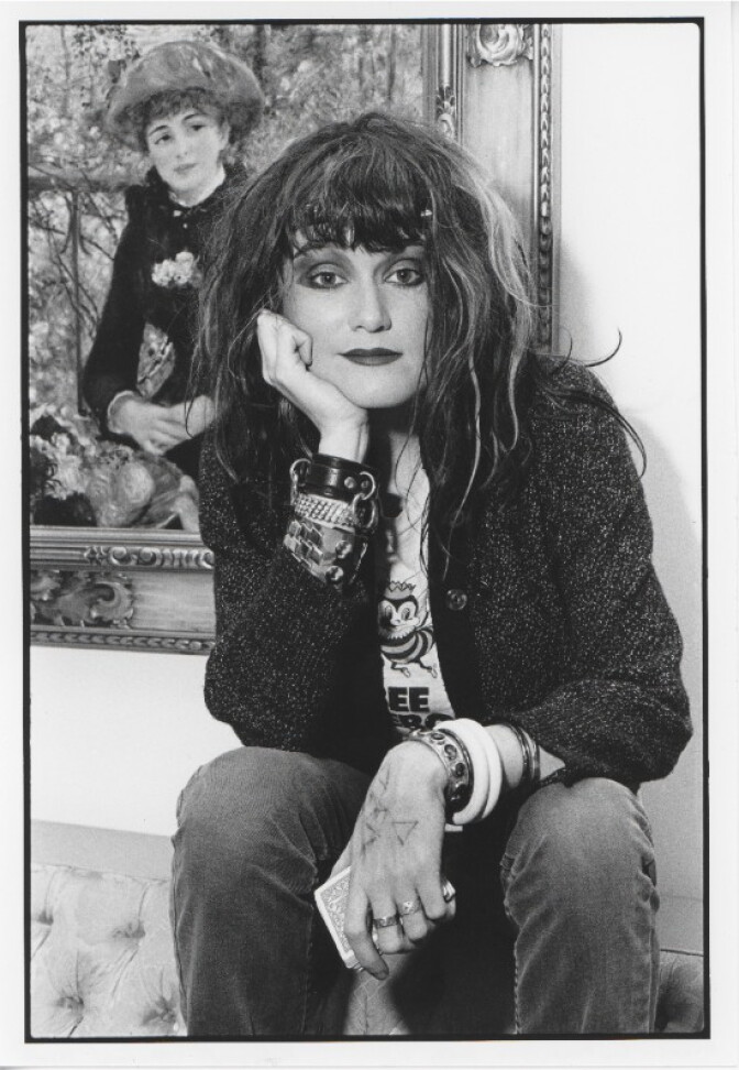 A person with tousled hair rests their chin on their hand, wearing bracelets and a bee-print shirt, seated before a framed painting.