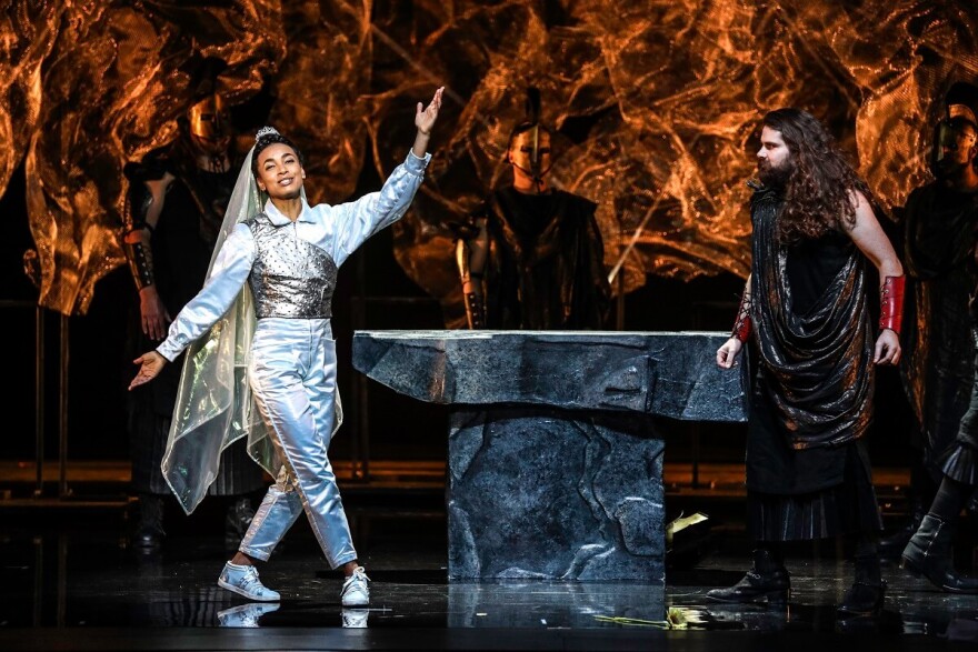 A woman in a veil and a sparkling pantsuit is joined by two men dressed as Greek gods on a stage around a stone table.