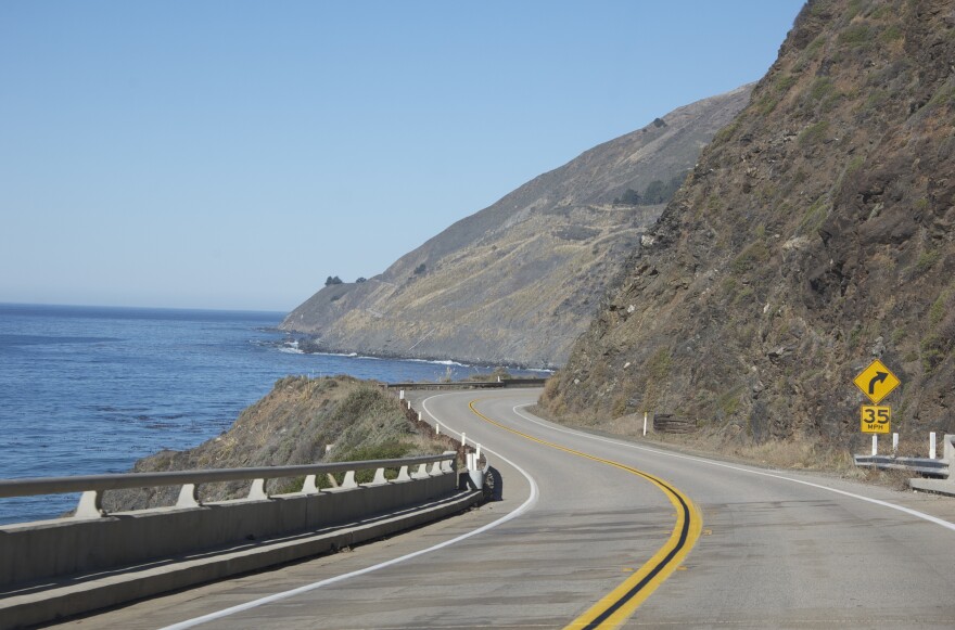 A windy roadway next to the ocean, on the right it's surrounded by hills. 