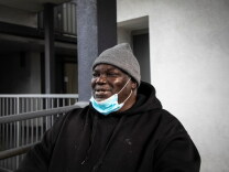 A middle-aged Black man wearing a black hoodie, grey knit hat and blue face mask smiles while looking off to the left. He is on what appears to be an outdoor walkway at a grey stucco apartment building. 