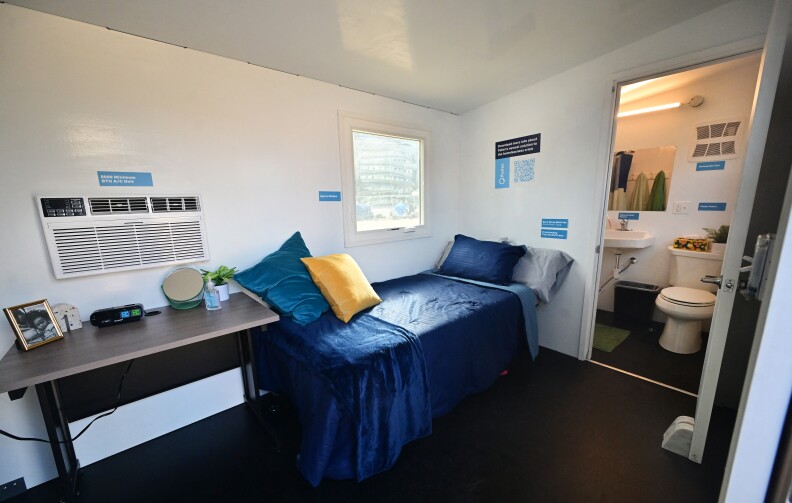 A single bed has blue sheets and pillows in a narrow space. A wall AC and window are on the same side of room as bed. There's an open door to a small restroom.