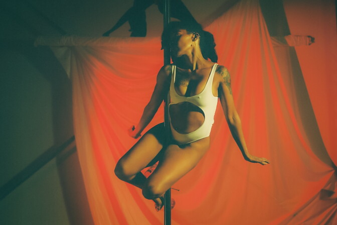 A feminine presenting person with medium-dark skin tone wears black curly hair to the side and a one piece white leotard with the belly cut out while balancing on a pole with their arms to the side. Behind them is a red curtain. 