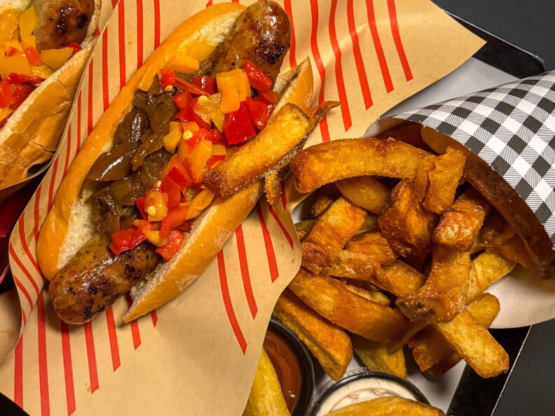 A German sausage with onion and pepper topping next to thick-cut French fries. 
