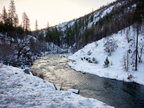 A stream cuts through a valley with snow-covered hills and pine trees on both sides.