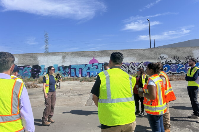 People in high-visibility safety vests stand near a graffiti-covered concrete wall outdoors.