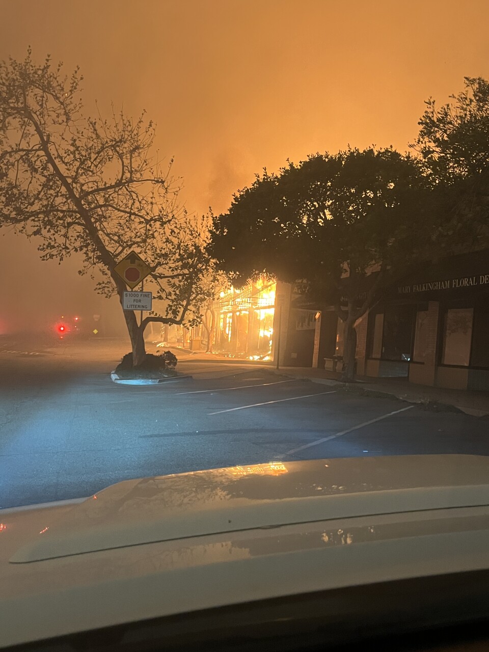Flames engulf a storefront at night, casting an orange glow through thick smoke as trees and empty parking spaces frame the burning building.