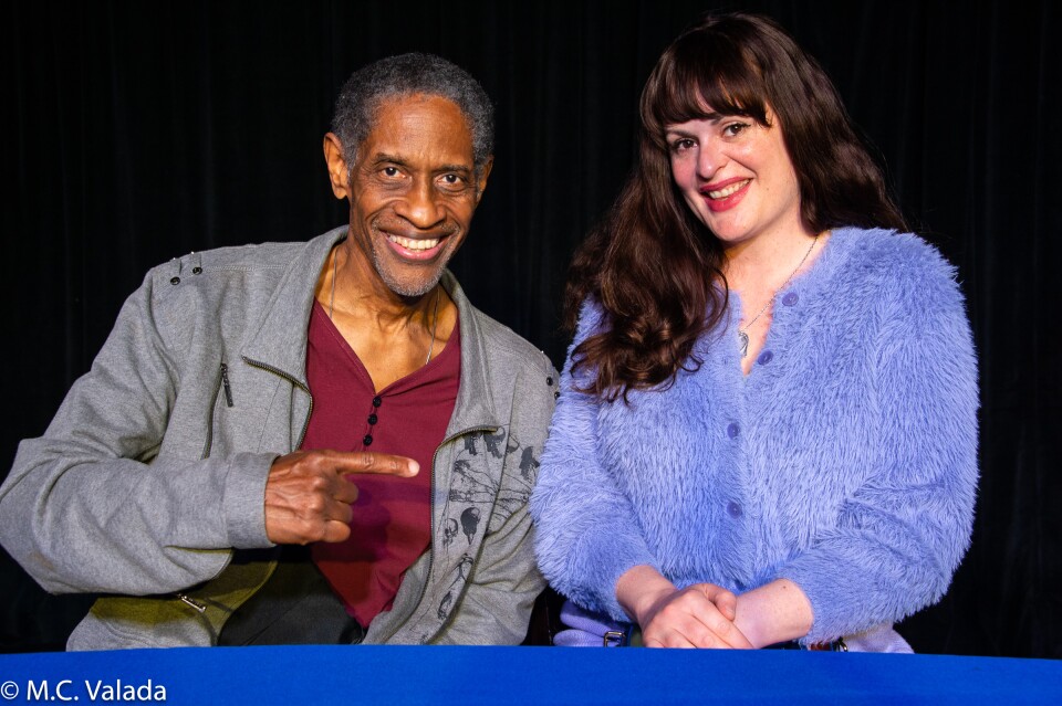 Tim Russ, a Black man with gray hair and beard and Amber Nelson, a white woman with long brown hair wearing a fuzzy purple sweater, smile at camera