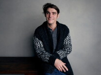 RJ Mitte, a smiling person with dark, tousled hair, sits against a gray studio wall, looking off to the side. They wear a black-and-white patterned knit cardigan over a gray henley.