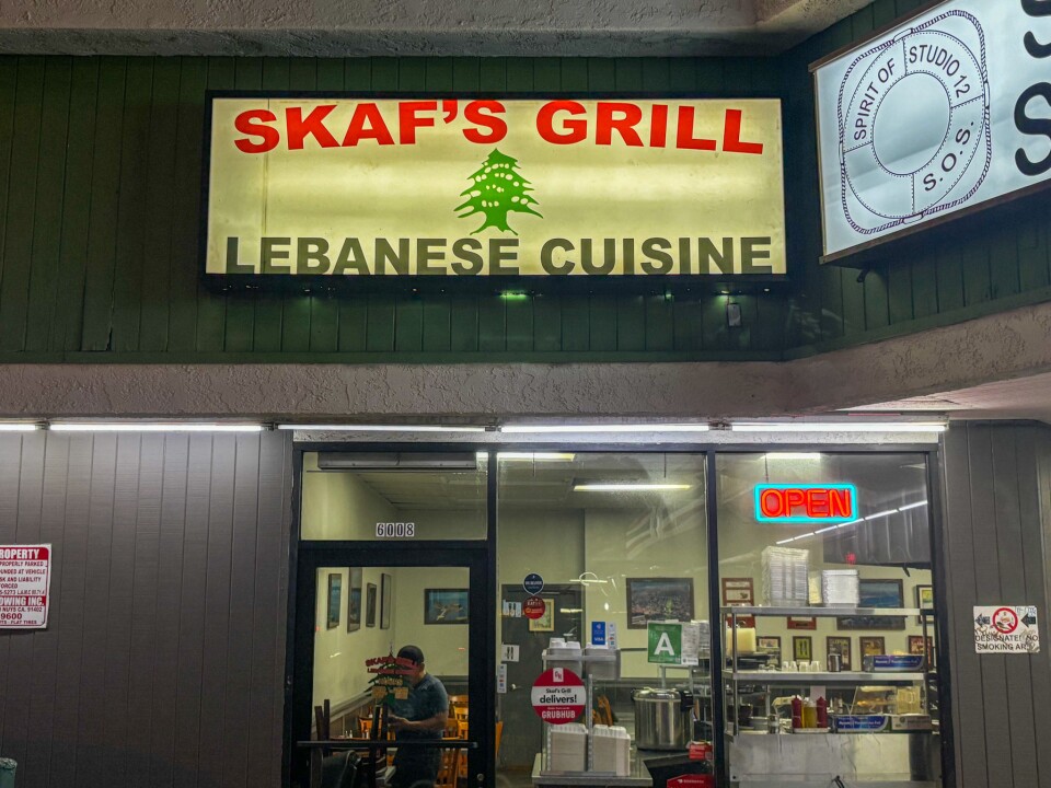 The exterior of Skaf’s Grill, a Lebanese restaurant with a bright sign reading “SKAF’S GRILL LEBANESE CUISINE” above the entrance. A neon “OPEN” sign glows in the window, with a glimpse inside showing framed wall art and a man seated at a table.