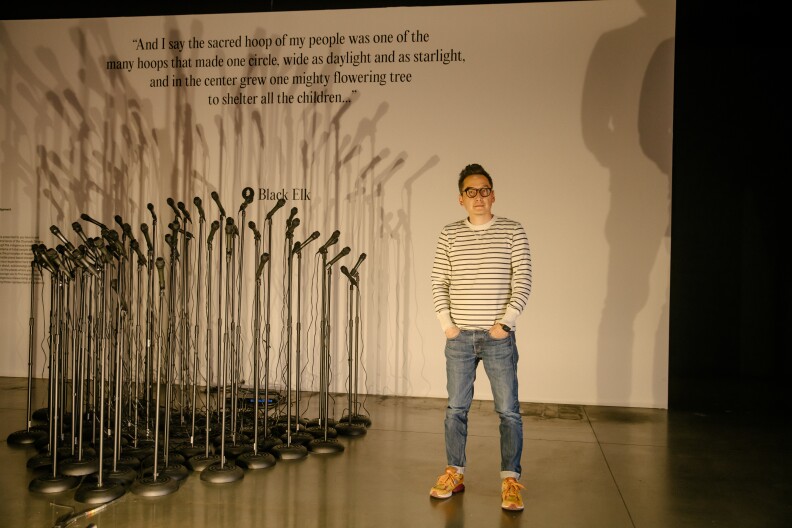 A man in glasses, a striped top, jeans, and sneakers stands in front of a collection of dozens of microphones on microphone stands, arranged in a tight triangle. He stands in front of a white backdrop with a quote on it: "And I say the sacred hoop of my people was one of the many hoops that made one circle, wide as daylight and as starlight, and in the center grew one mighty flowering tree to shelter all the children..." - Black Elk. Smaller type to the left explains what the exhibition is.
