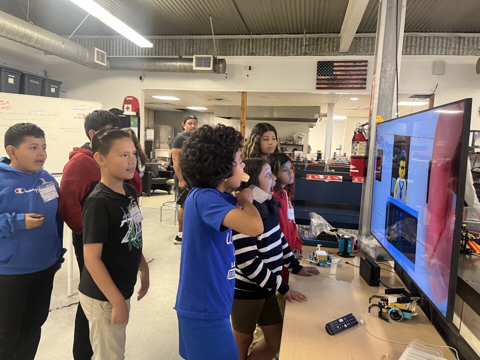 A group of kids stand around a large television screen at a local youth tech center.
